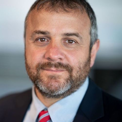 Portrait of a middle-aged man with short brown hair and a salt-and-pepper beard, wearing a suit and red striped tie, looking directly at the camera.